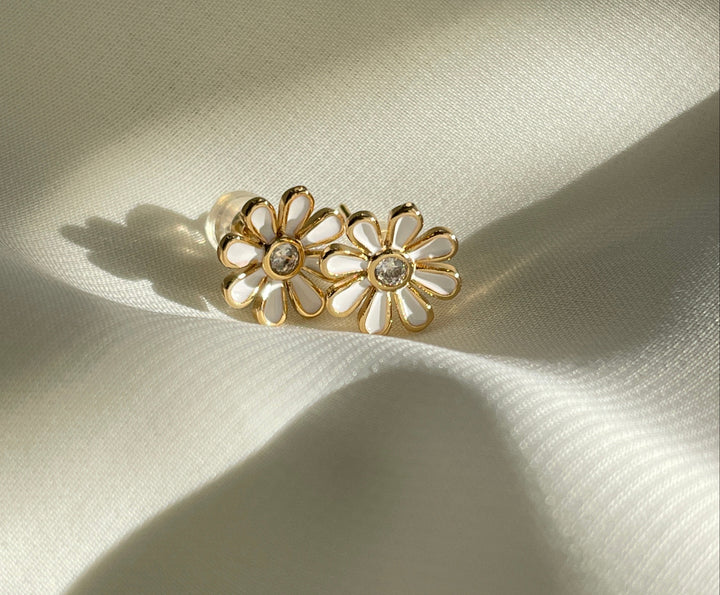A pair of white enamel daisy post earrings with gold plating, displayed on a textured beige surface.